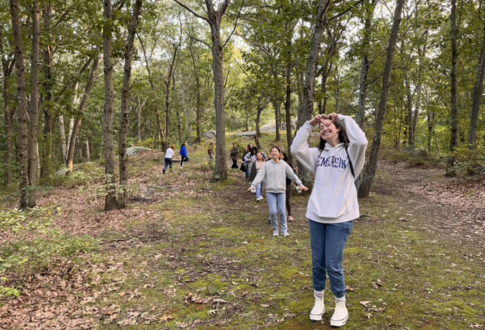 students walk in woods, student in front makes heart shape with hands