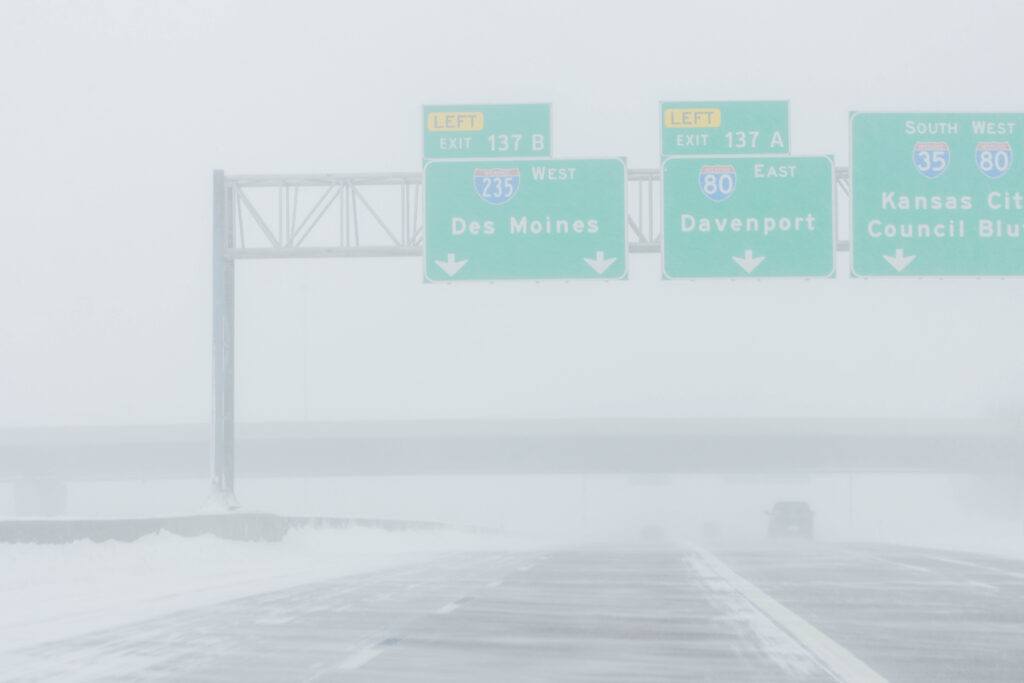 A snowy highway with three large signs suspended over the road, and only one car on the road