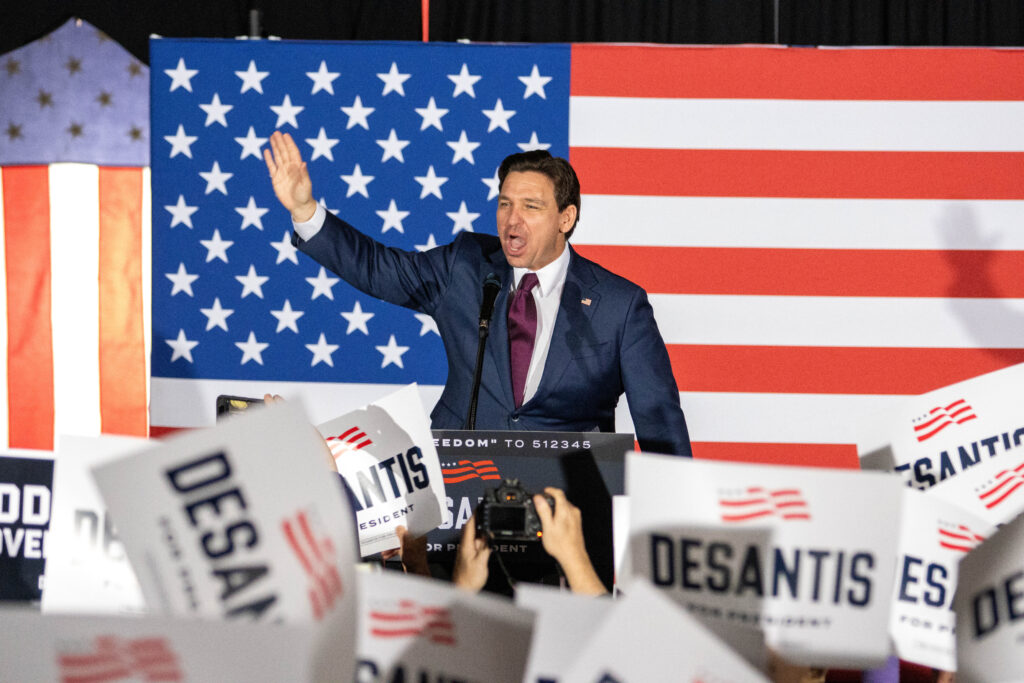 Ron DeSantis raises his hand while on stage in front of a lectern on stage in front of American flag