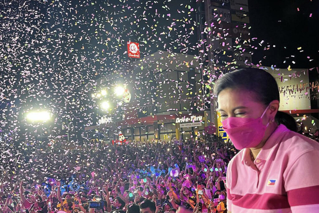 woman in pink shirt, pink mask stands before crowd showered with confetti