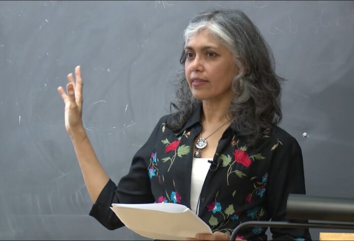 A woman stands in front of chalkboard, behind a podium, gesturing out to her audience.