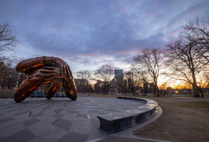A sculpture of arms embracing is in the forefront as the sun sets in the background.