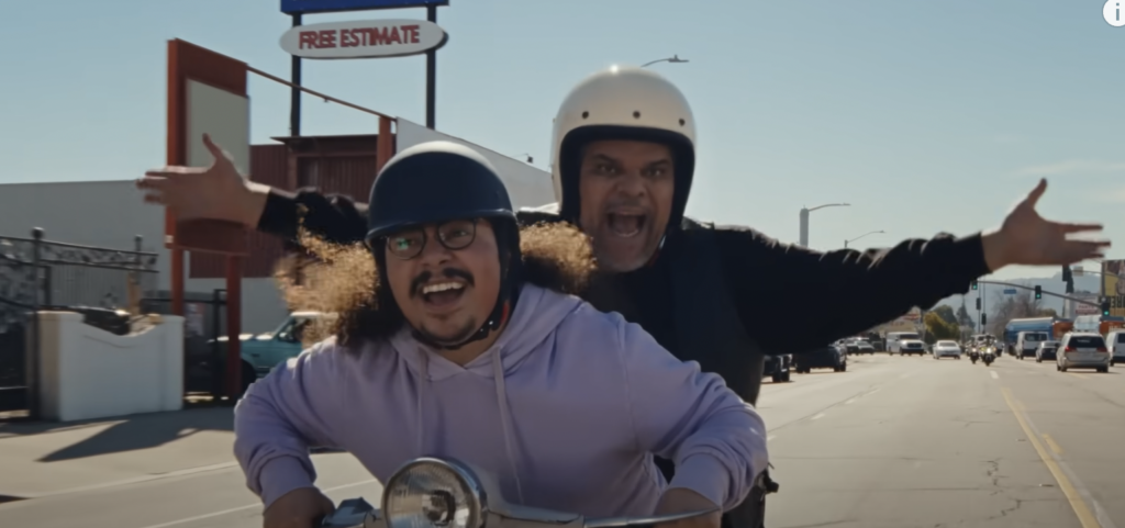 Luis Guzmán rides on the back of a scooter with Andrew Santiago in a T-Mobile ad.