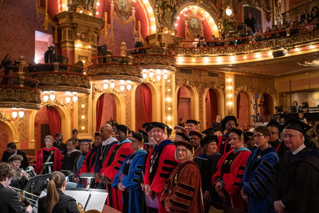 men and women stand in ornate theater wearing colorful regalia; members of orchestra in foreground.