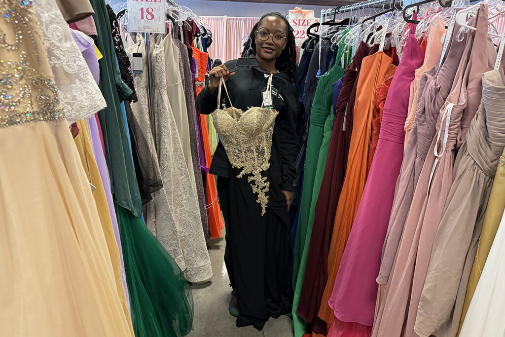Young Black woman holds a black gown with gold bodice between two rows of colorful gowns