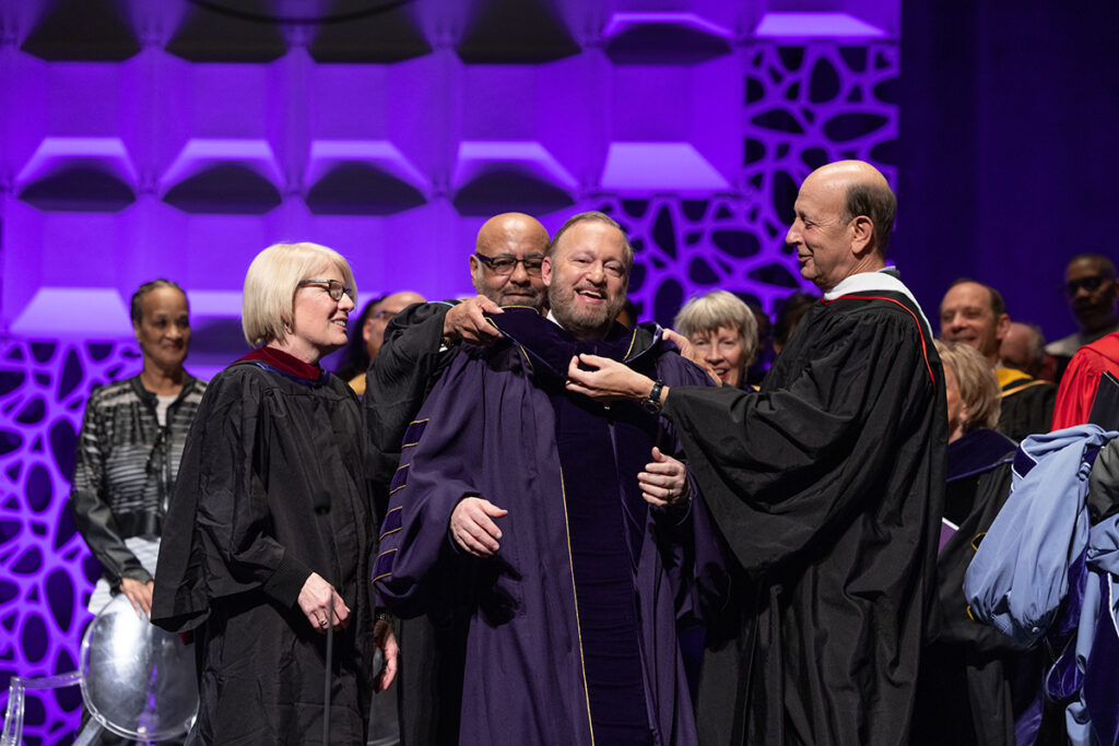 two men in black robes place academic hood over the shoulders of man in purple robes