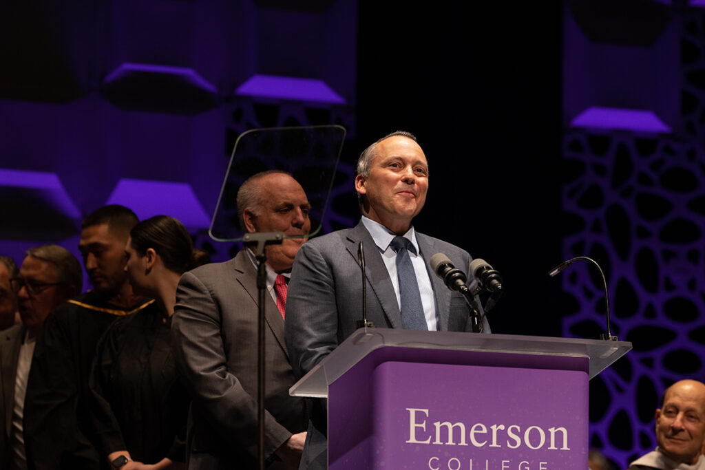 man in grey suit stands at podium, line of people behind him