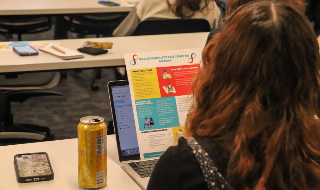 The back of a panel attendee is shown staring at a colorful handout. The handout is a resource on autism and the prevalence of neurodivergency nationally and worldwide.