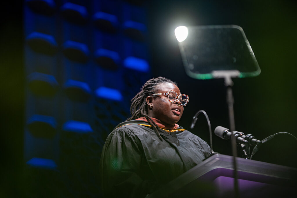 Woman in black robe, braided hair, colorful glasses reads at podium