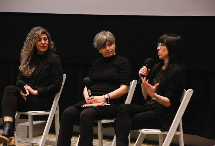 Three women dressed in black sit in white chairs against black background. One speaks into microphone