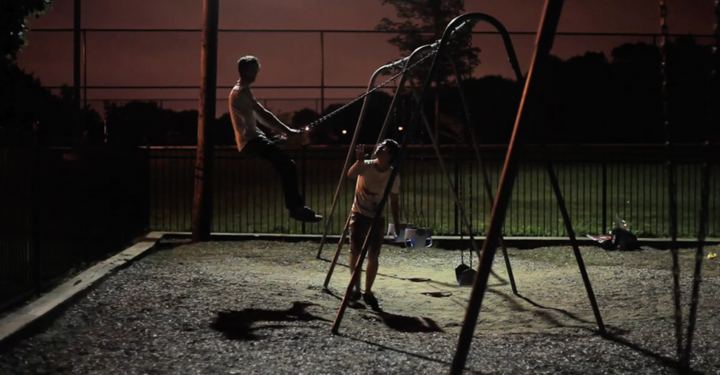 A person swings on a swing at night with another person standing next to them