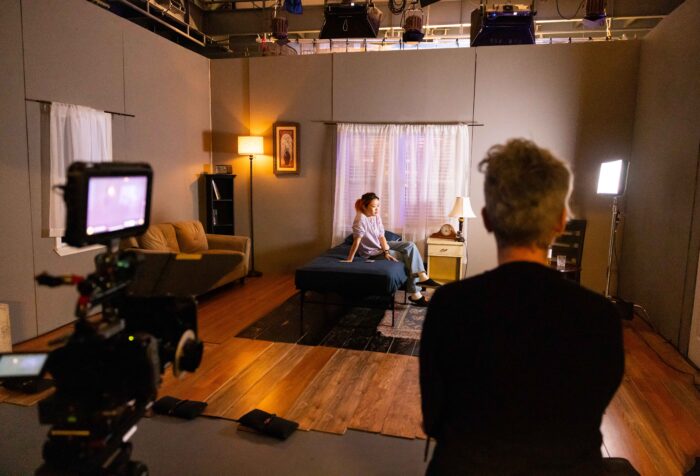 An actor sits on a bed on a soundstage during a filming. The director and film camera are in the foreground.