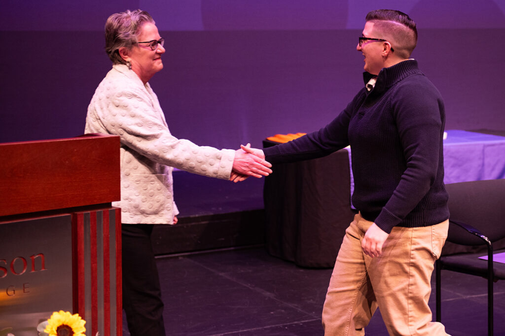woman with short grey hair, wearing glasses and a white jacket, shakes hands with faculty member crossing the stage wearing a navy blue sweater, glasses, and a military style haircut.