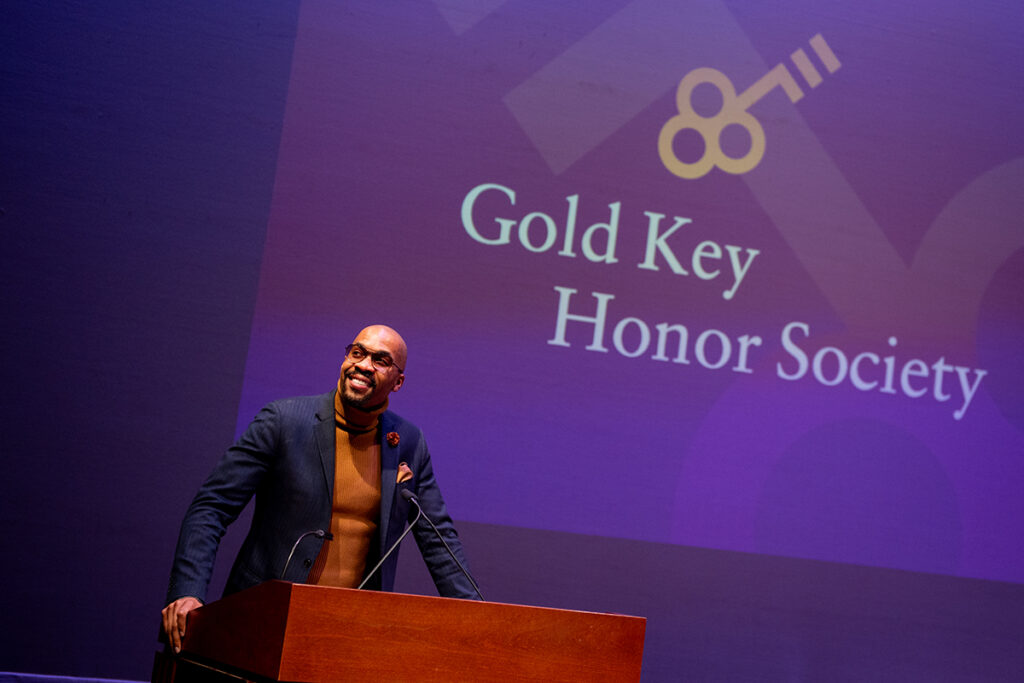 African American man wearing orange turtleneck, navy jacket, glasses smiles at podium, words Gold Key Honor Society projected behind him
