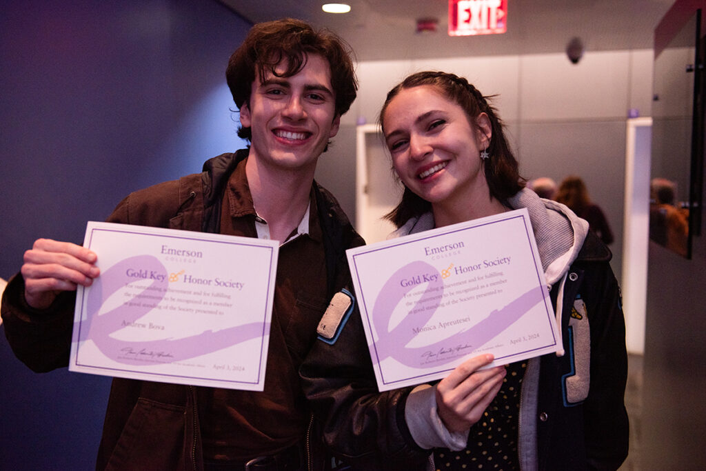 Two students wearing jackets hold up certificates featuring purple Emerson flourish