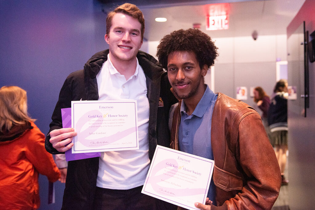 Two students, one white, one of color, hold certificates in a hallway