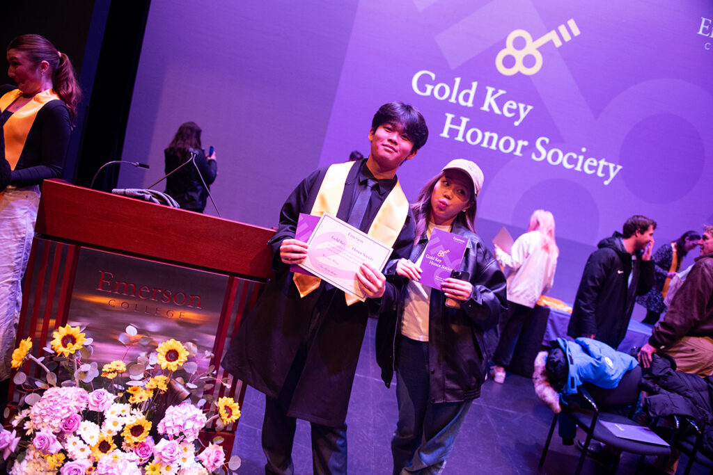 Student wearing gold academic sash holds certificate as friend stands next to him holding ceremony program. The students stand next to a podium reading Emerson College behind a bouquet of flowers