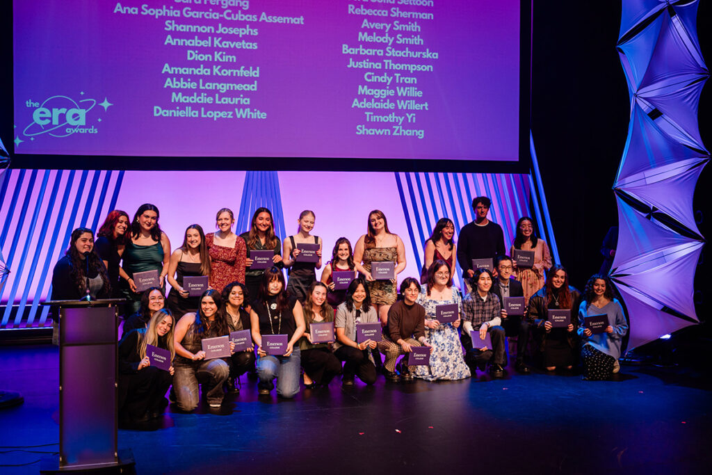 Students gather on stage holding purple certificate cases, screen listing names behind them