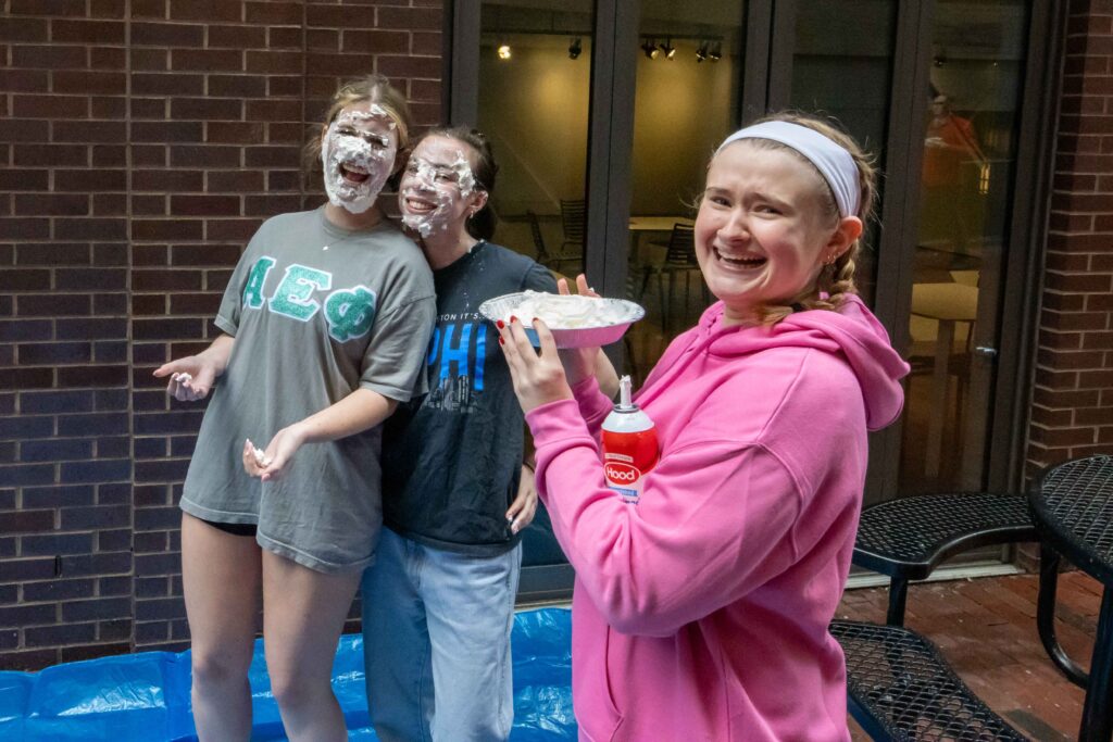 Phoebe Alves with pie on her face, Ashley Bradshaw, and Emma Weiss holding a pie and smiling