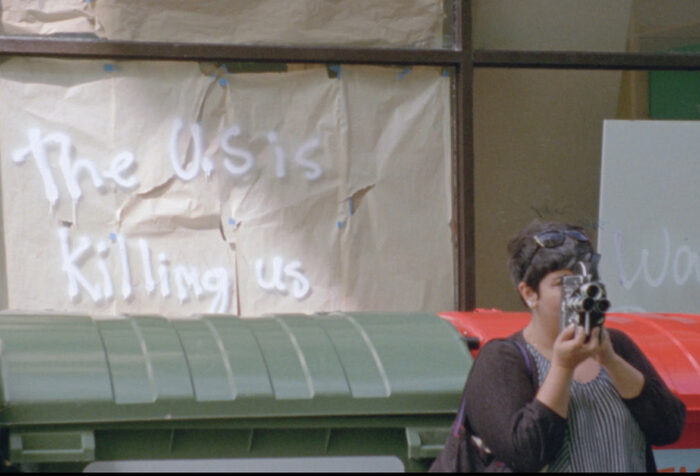 Person holding video recorder to their face with the text 'The US is Killing us' written on a wall behind them
