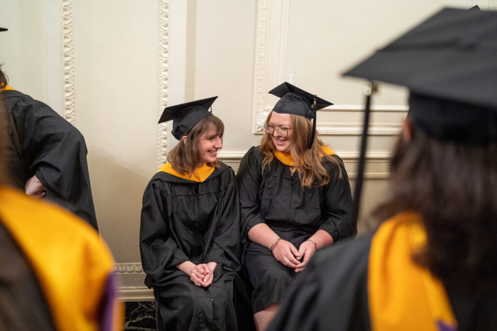 Two grads sit next to each other smiling and talking