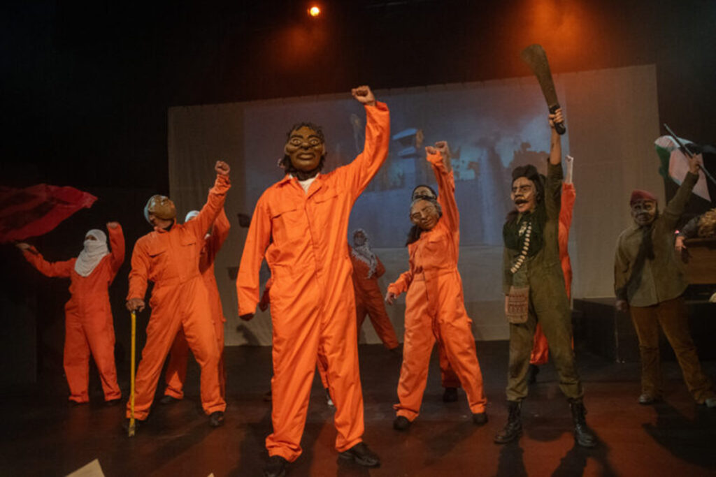 figures with papier mache heads wearing orange prison jumpsuits and green fatigues stand with fists raised
