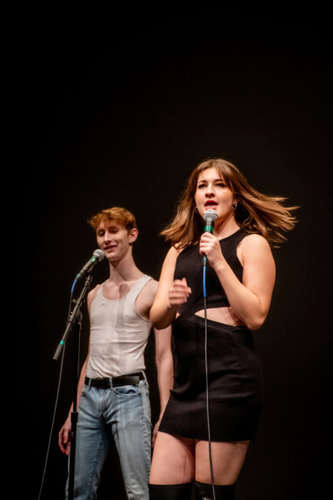 Two students, one in white tank top, jeans, with short red hair, one in black tank dress with long brown hair, sing at microphones