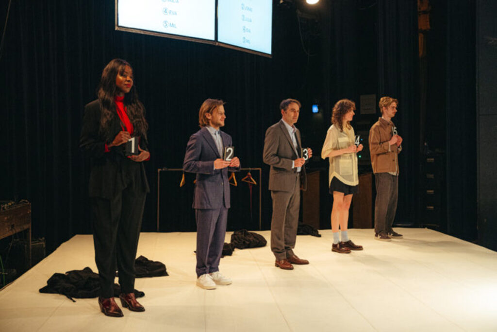Five people stand on stage holding black cards numbered 1 to 5.