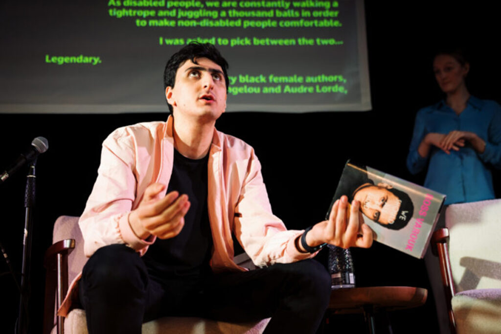Seated man in pink jacket looks up, holding a book in his left hand. Screen behind him has green words projected onto it