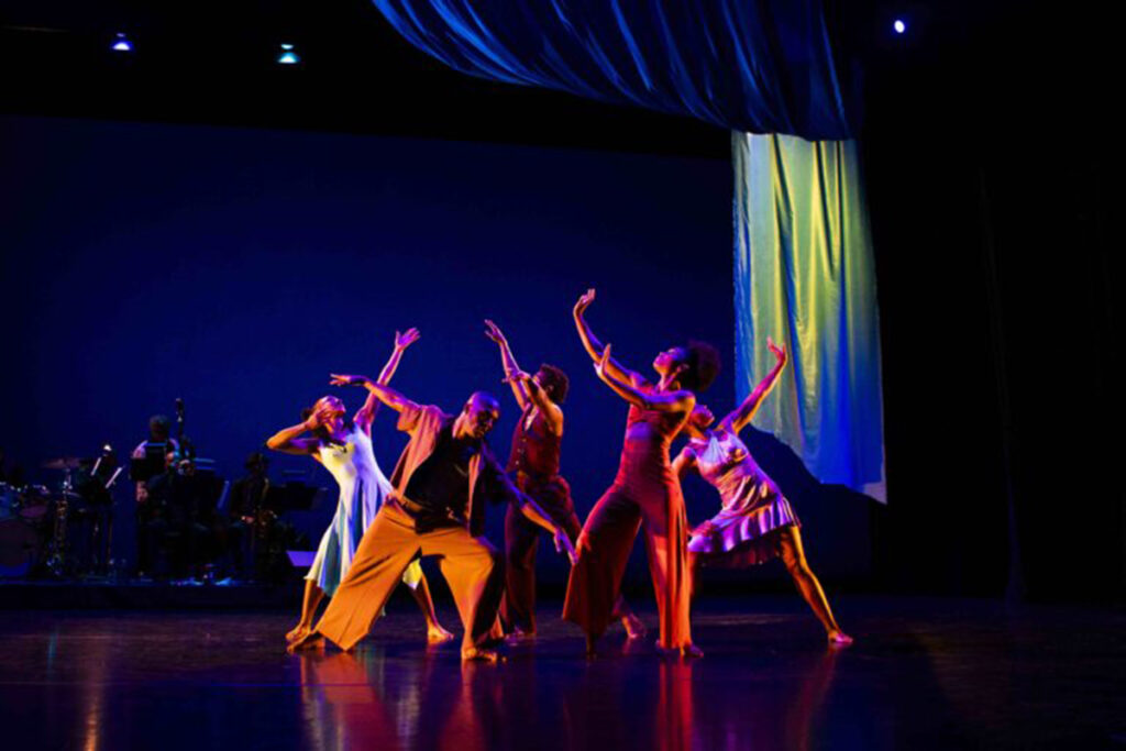 Black dancers pose in a tight formation on stage, musicians in silhouette behind them