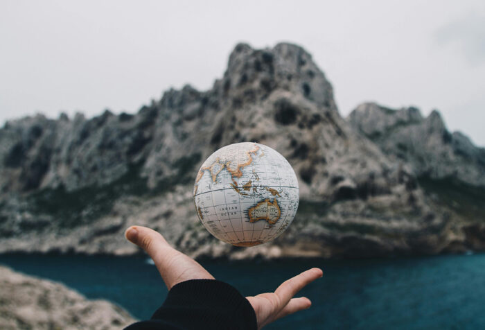 hand tossing small globe in front of a rocky outcrop over deep blue water