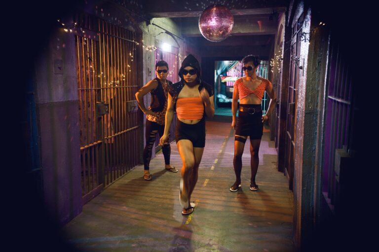 Three men in orange crop tops, tight shorts or leggings and sunglasses dance between prison cells, a glitterball hanging above them