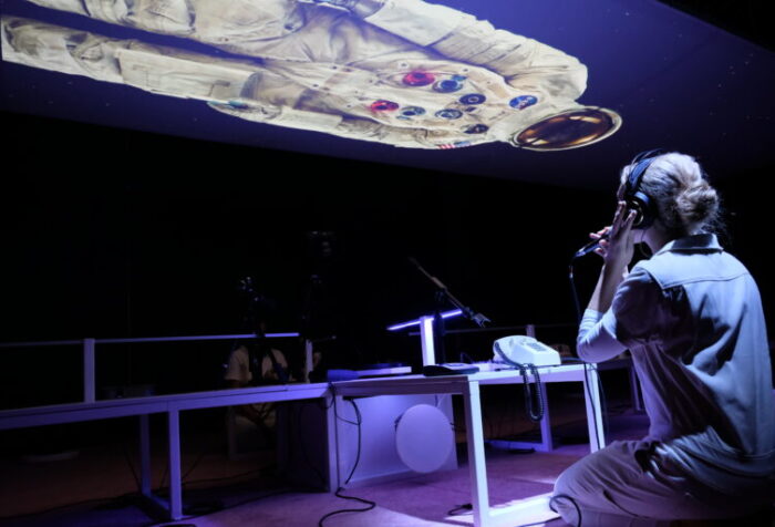 person wearing headphones kneels at desk with a telephone on top and speaks into a microphone; image of astronaut is projected on ceiling