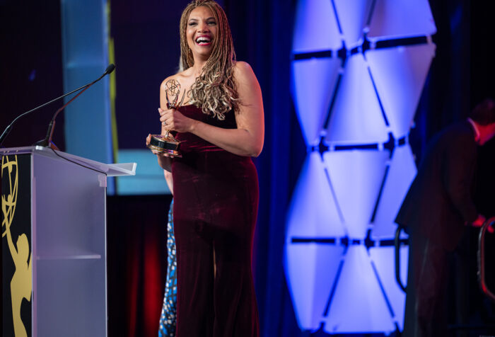 woman with long hair in braides, wearing a burgundy velvet strapless gown smiles and holds Emmy next to podium