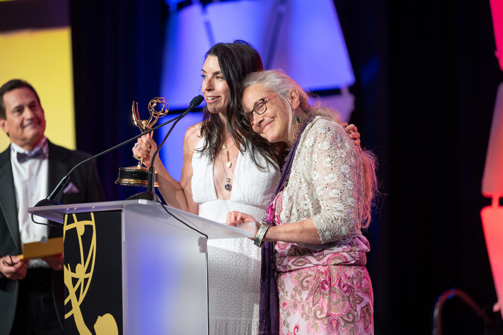woman with long, dark hair in white sleeveless dress, heart-shaped necklaces, holding Emmy stands at podium with woman with long grey hair, glasses, pink paisley dress