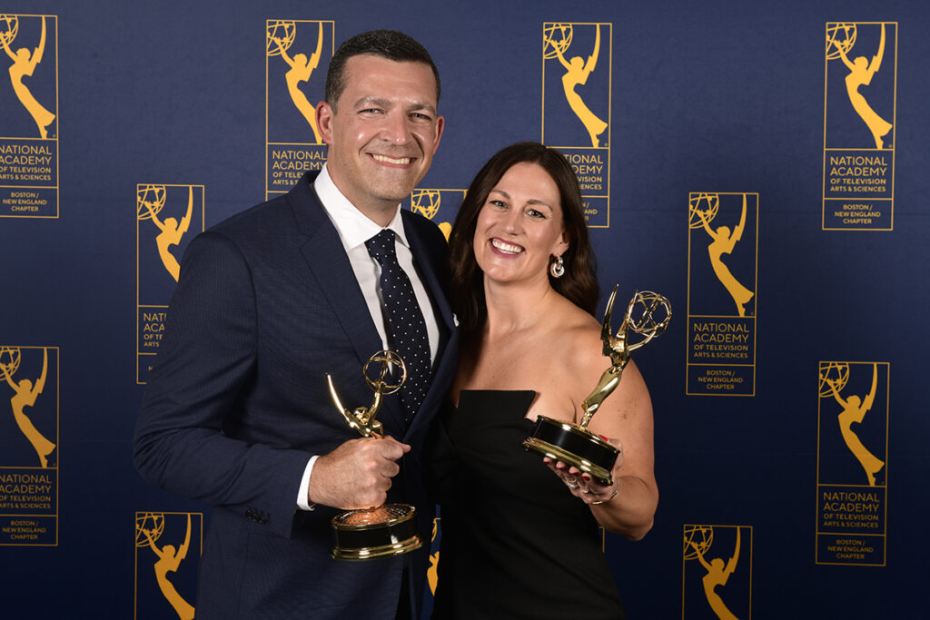 Man in navy blue suit, navy polka dot tie, and woman in black strapless dress hold Emmys