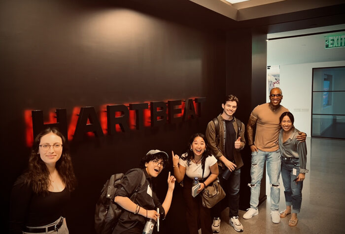 Emerson students and faculty with a HartBeat Productions employee pose in front of a wall sign that says HartBeat