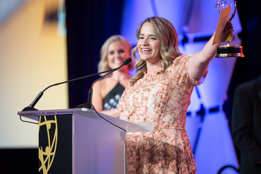 woman with long blonde hair, wearing a pink floral dress, holds up an Emmy while standing at a podium