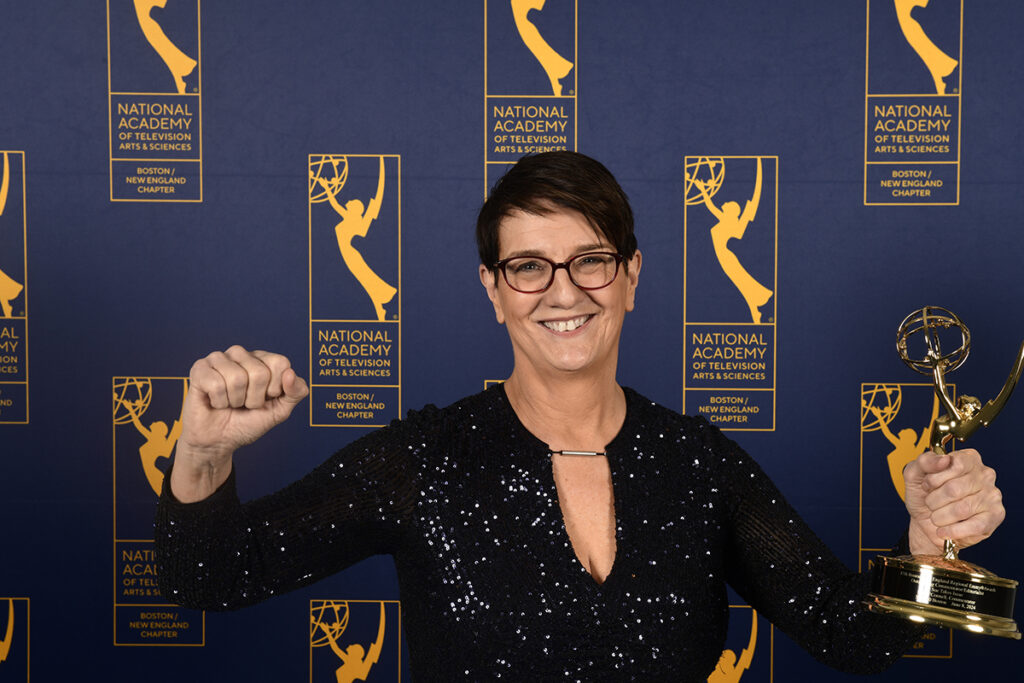 Woman with short dark hair, glasses, in black sequined dress, pumps fist and holds Emmy in front of a step and repeat