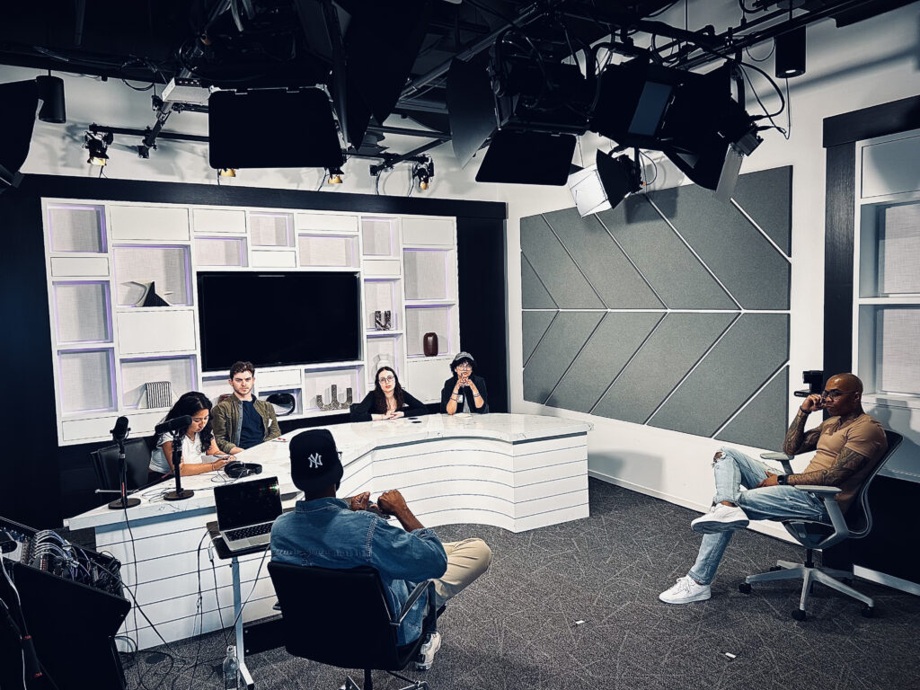 Four people sit a news desk listening to one person in a chair and another person sitting in a chair