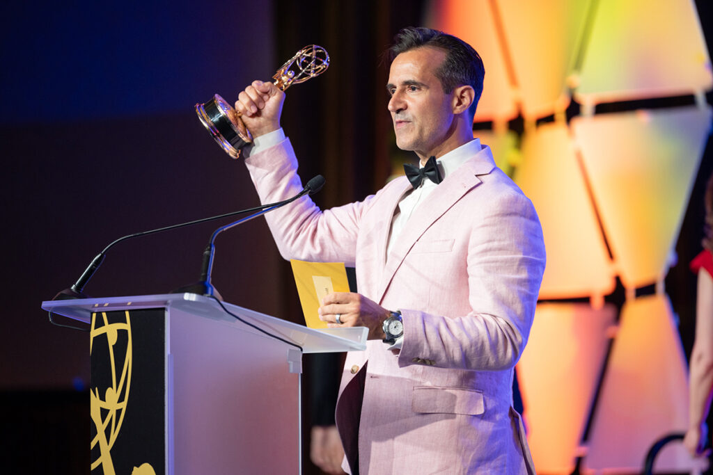 man in pink linen jacket, black bowtie, raises Emmy Award at podium