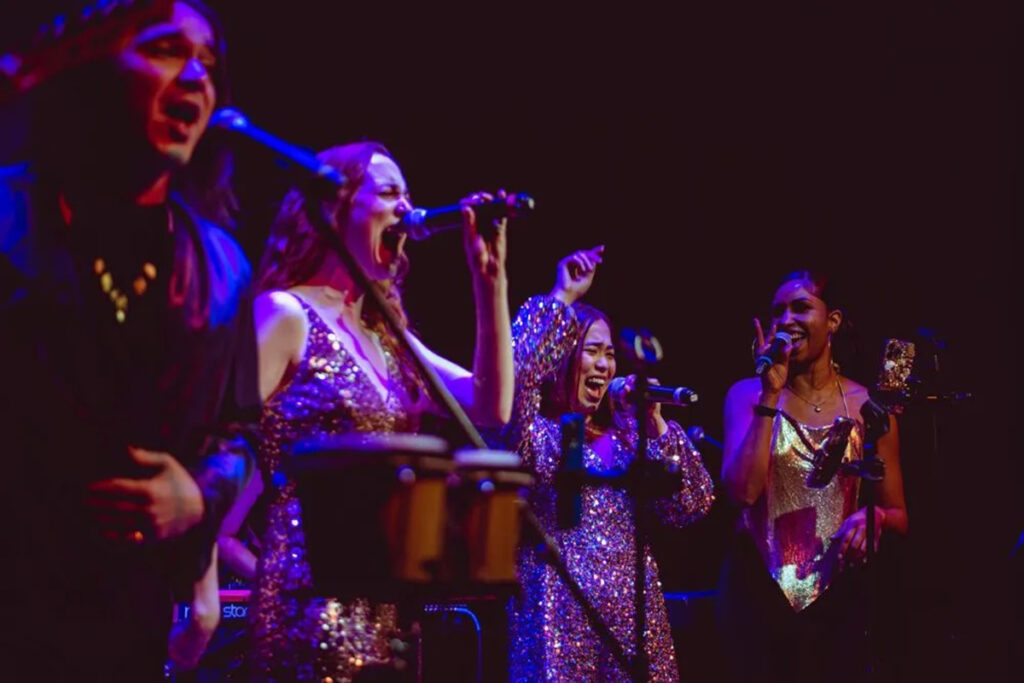 Four women and/or nonbinary musicians in sparkly tops sing into mics on a dark stage