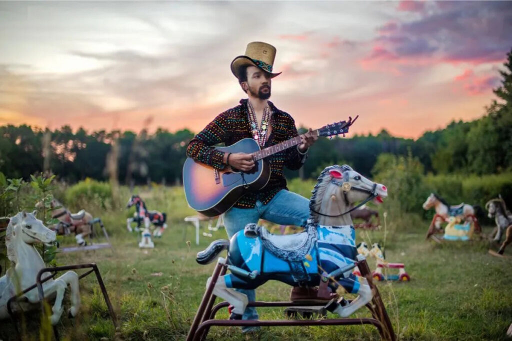 bearded white man in straw hat, patterned shirt, jeans, plays guitar in a field surrounded by rocking horses