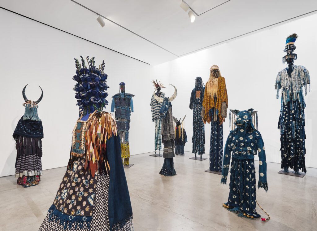 A group of sixteen characters clad in hand-dyed fabrics, woven details, and ornate stitching, many standing at the height of stilts