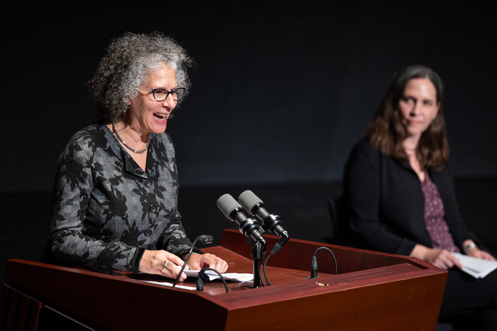 Woman with grey curly hair, wearing black framed glasses and black and grey shirt, speaks at podium