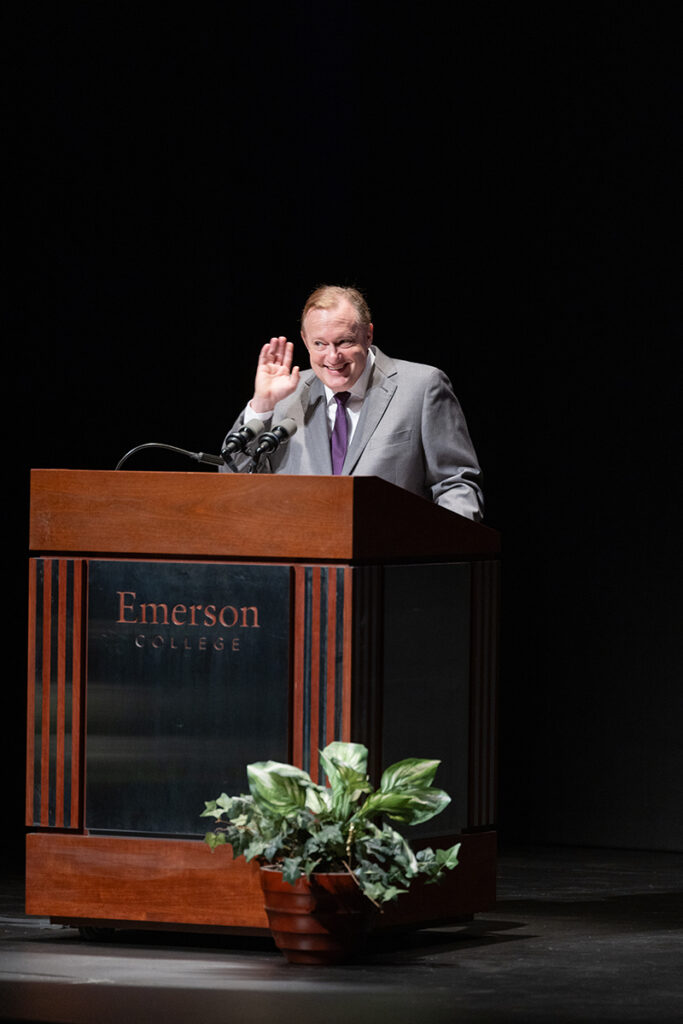 Man with short reddish hair, wearing grey jacket, purple tie, holds hand up to ear in listening gesture while standing at podium surrounded by plants