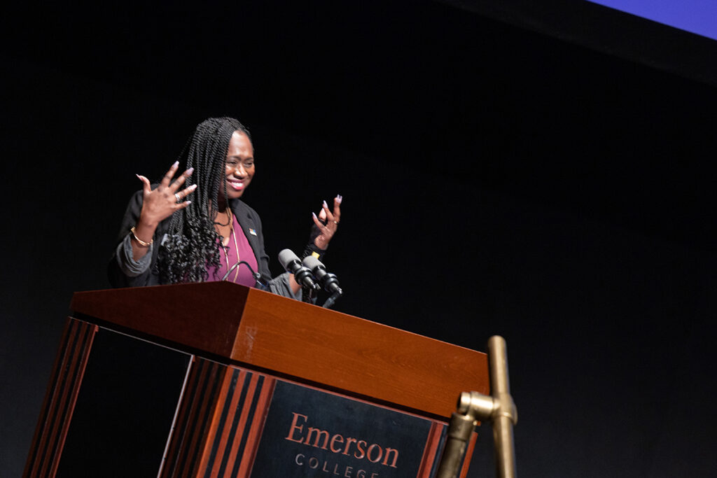 Woman with long hair in braids, wearing purple shirt, black jacket, gestures and smiles at podium