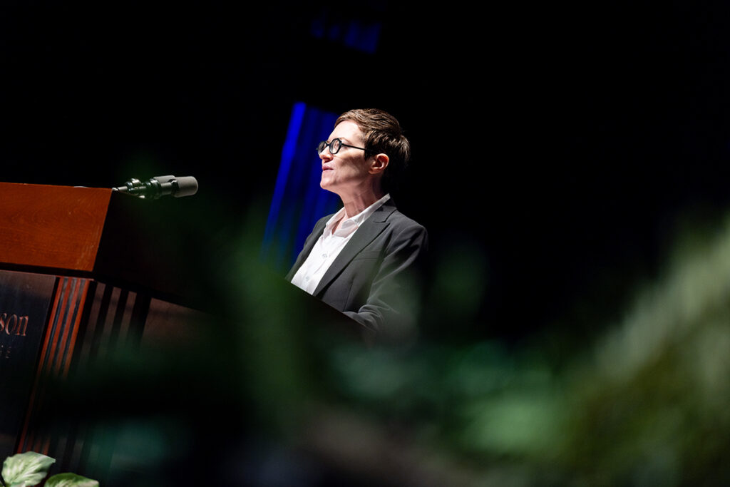 Woman with short brown hair wearing white shirt, black jacket, black round framed glasses speaks at a podium