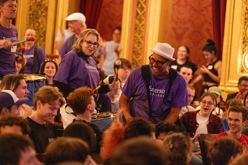 People wearing purple Emerson T-shirts play drums inside theater as audience smiles and claps