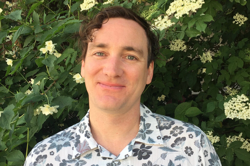 white man with curly brown hair wearing grey and white floral shirt stands in front of flowering plants